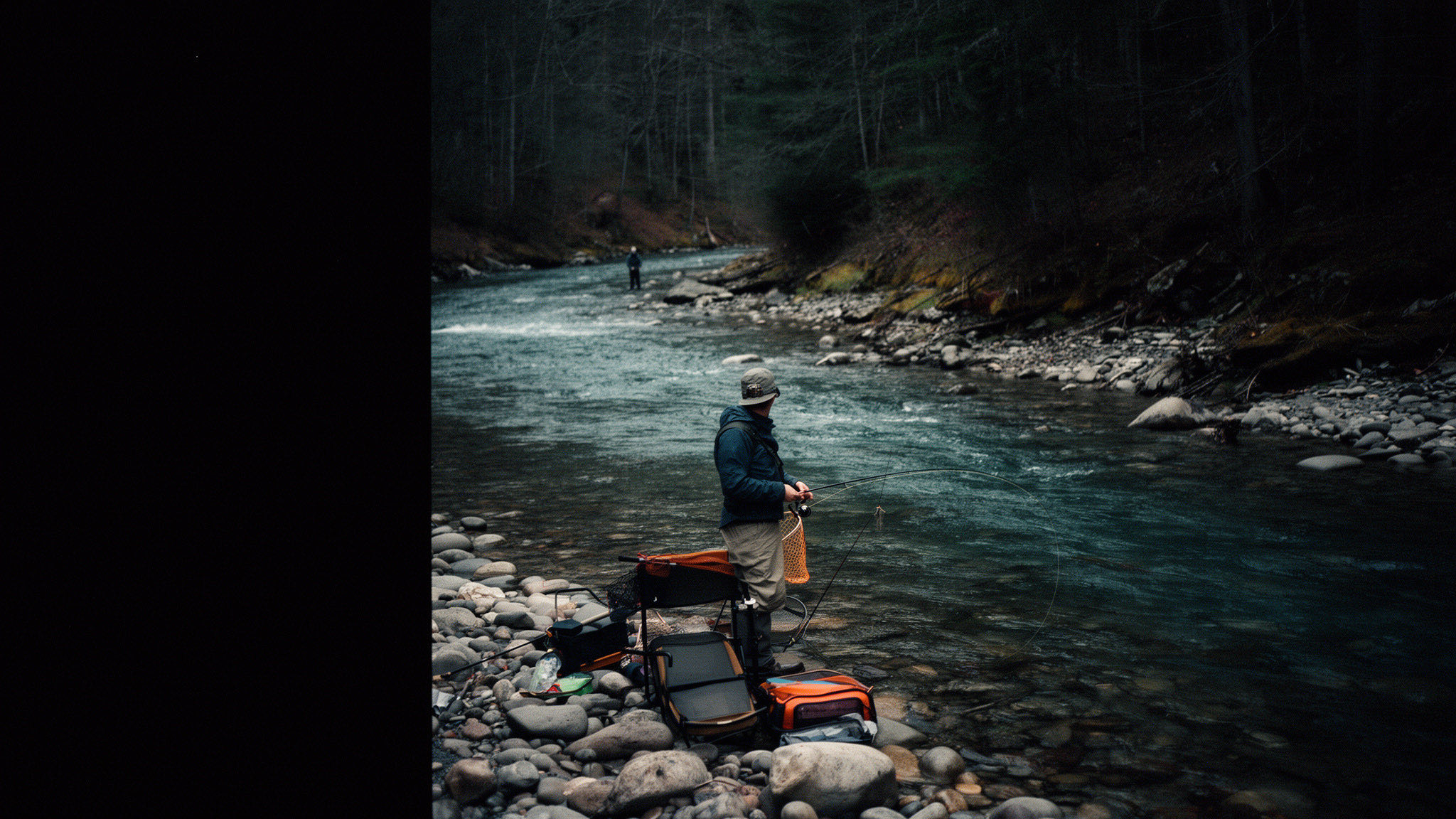 A fly angler stands beside a clear Appalachian river bordered by forest and rounded stones